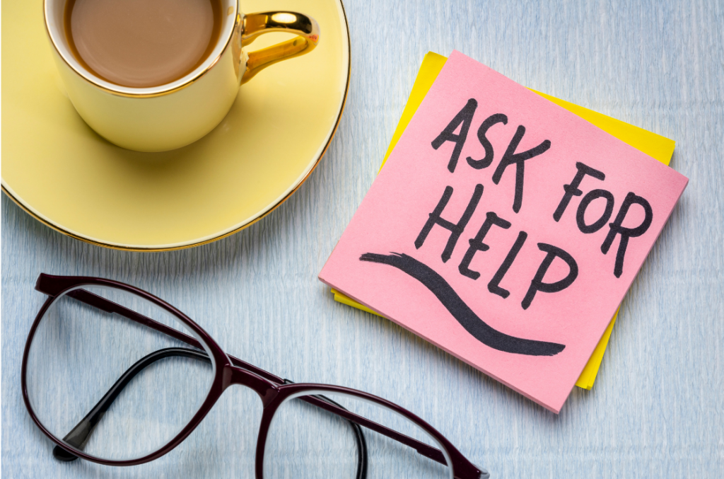 a pink post it not with "ask for help" next to a yellow tea cup and saucer and glasses